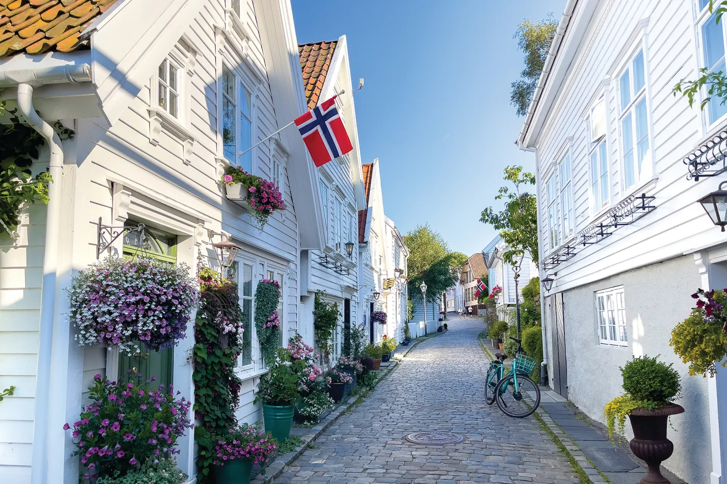 Cobblestone street with white wooden houses, flowers, a Norwegian flag, and a bicycle.