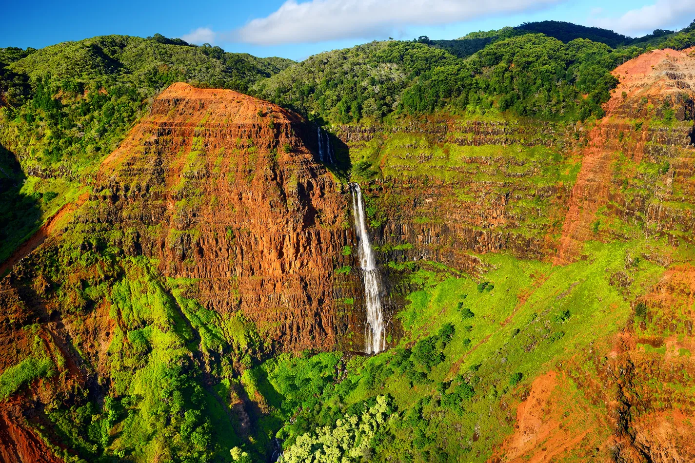 Tall waterfall cascading down the red and green cliffs of Waimea Canyon on Kauai beneath a bright, sunny sky.